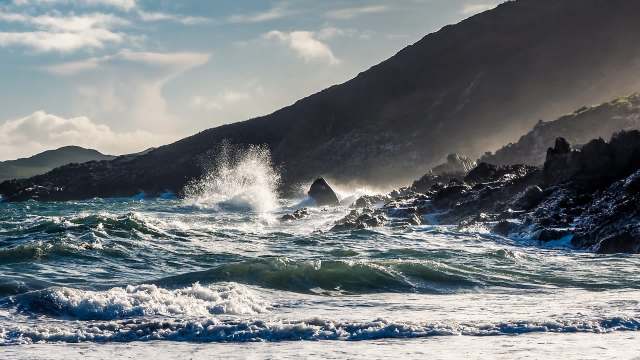Waves crash against rocky shoreline with mountains in the background under a partly cloudy sky.