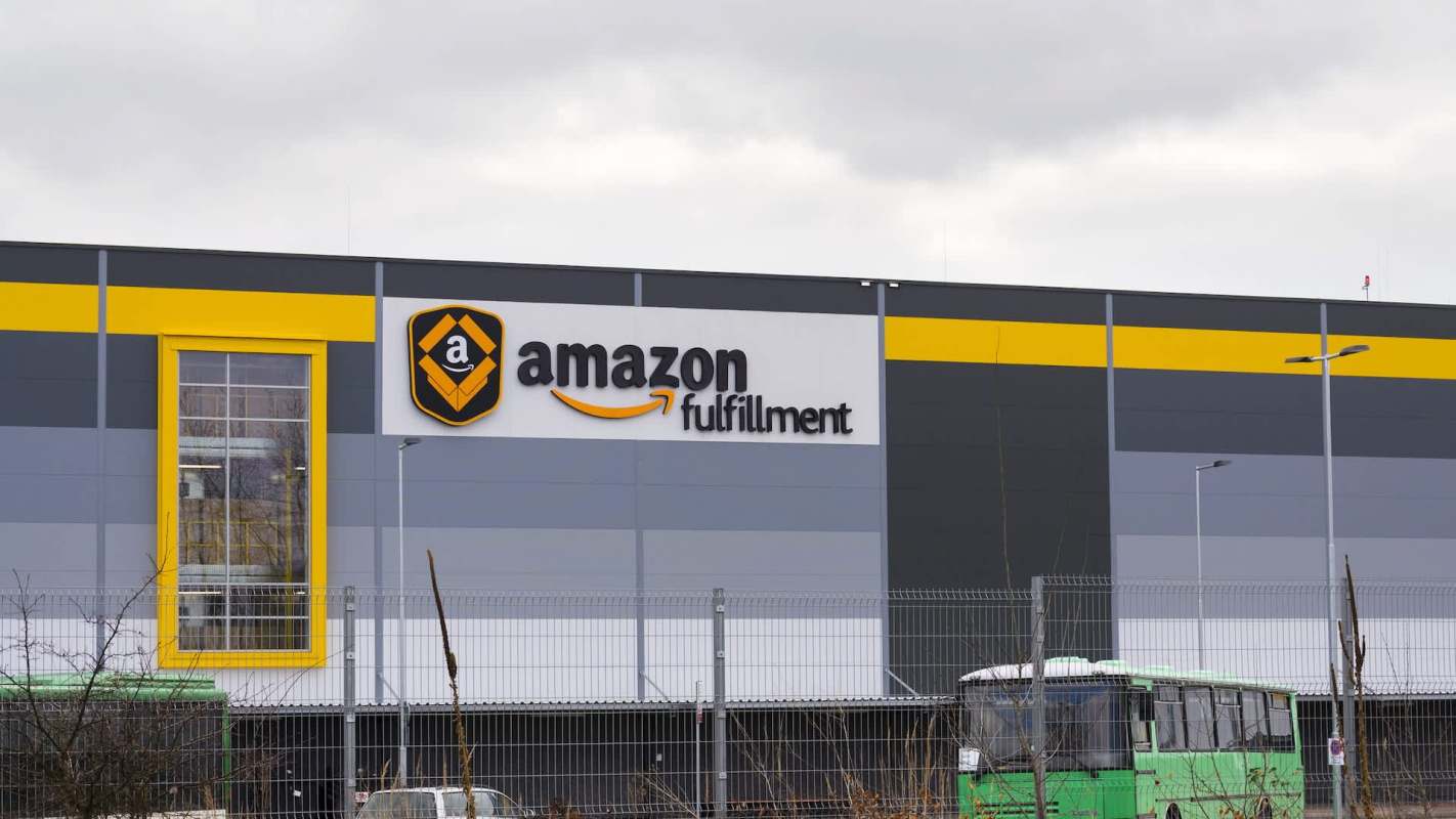An Amazon fulfillment center with a prominent sign and a green bus parked nearby.