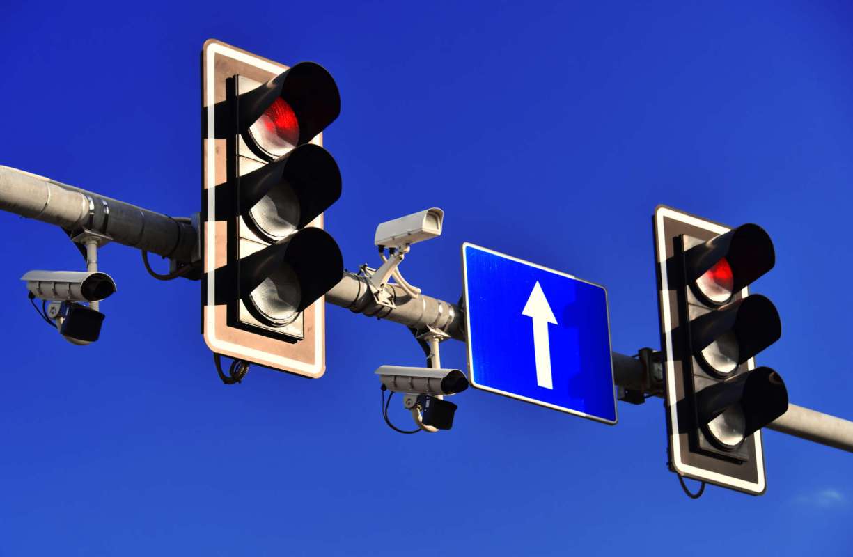 Traffic lights displaying red signals with a road sign indicating straight ahead, under a clear blue sky.