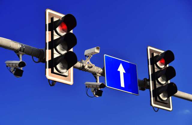 Traffic lights displaying red signals with a road sign indicating straight ahead, under a clear blue sky.