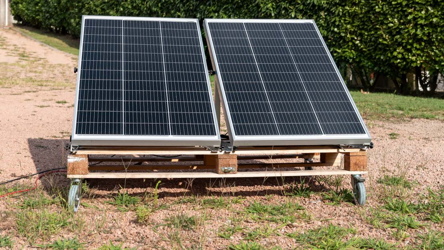 Two solar panels mounted on a wooden frame with wheels, placed on gravel ground.