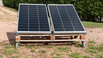 Two solar panels mounted on a wooden frame with wheels, placed on gravel ground.