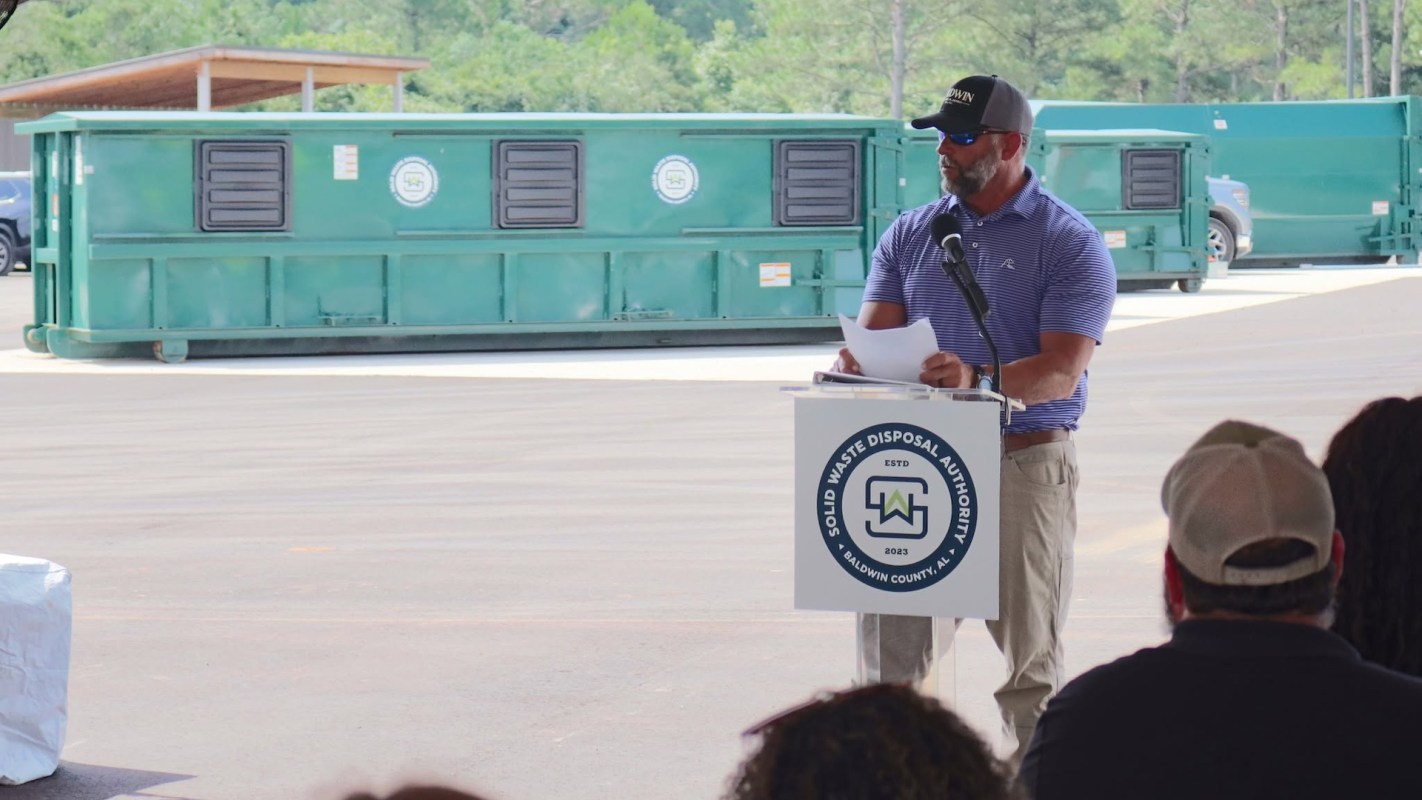 A speaker presents at an outdoor event near green waste disposal bins.