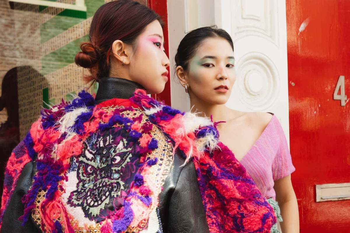 Two women stand against a red door, one wearing a colorful embellished jacket and the other in a pink top.