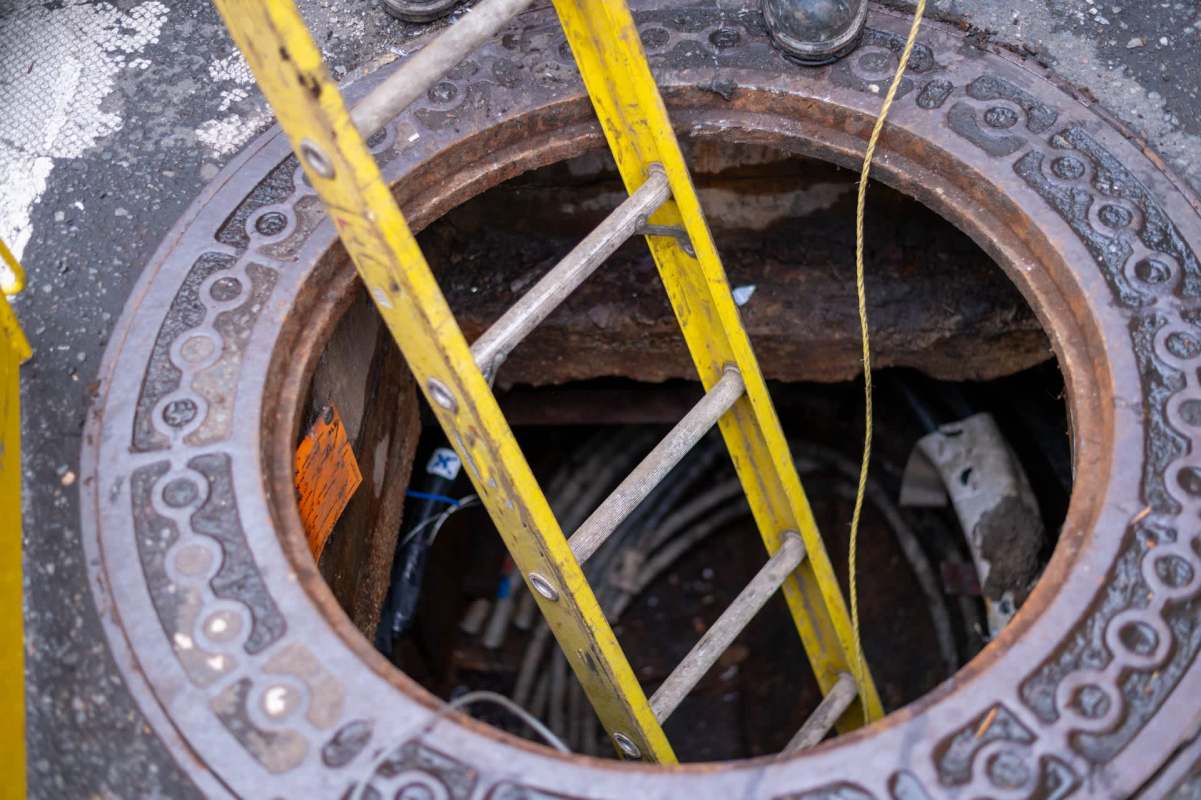 A yellow ladder leads into a round manhole with visible cables inside.