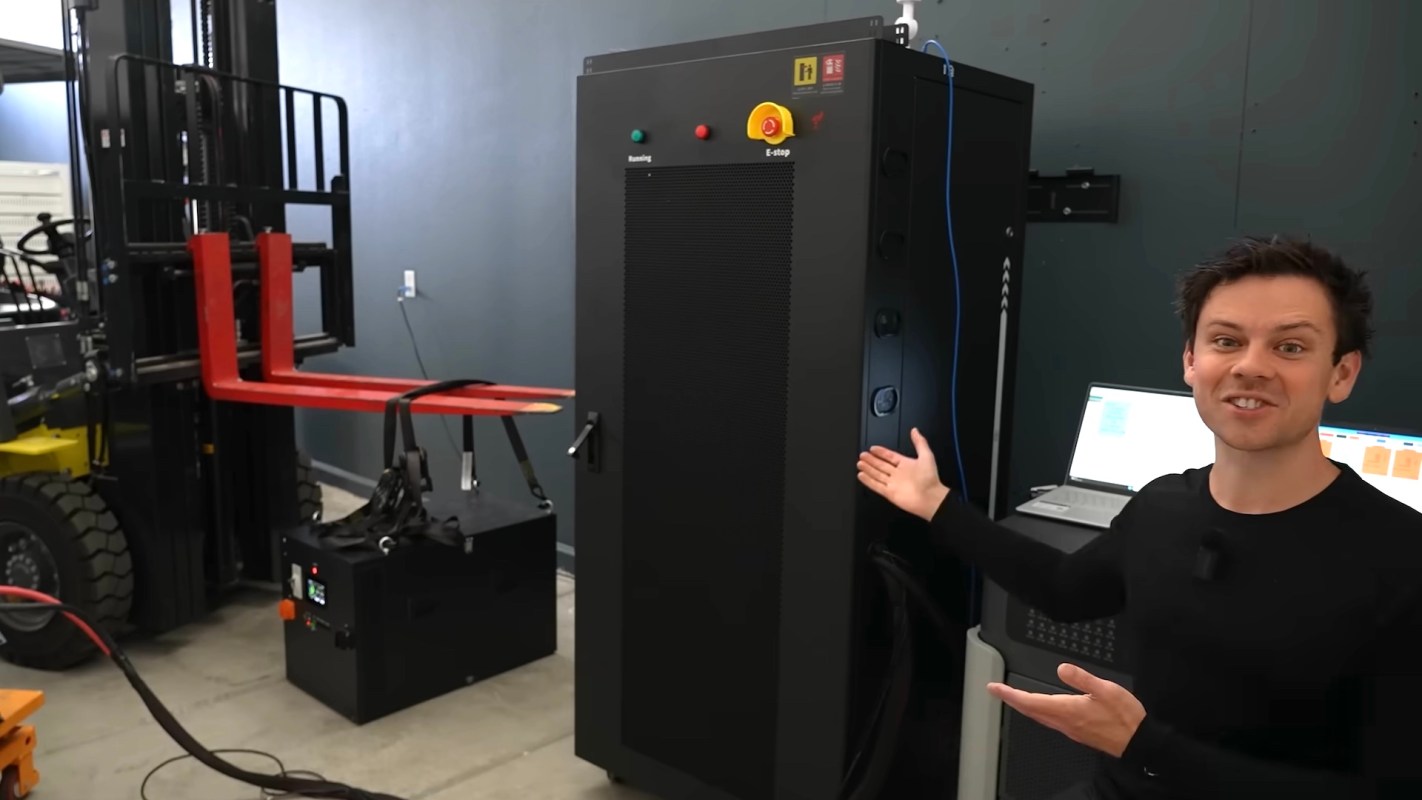 A person gestures towards a large black machine in a workshop with a laptop and equipment nearby.