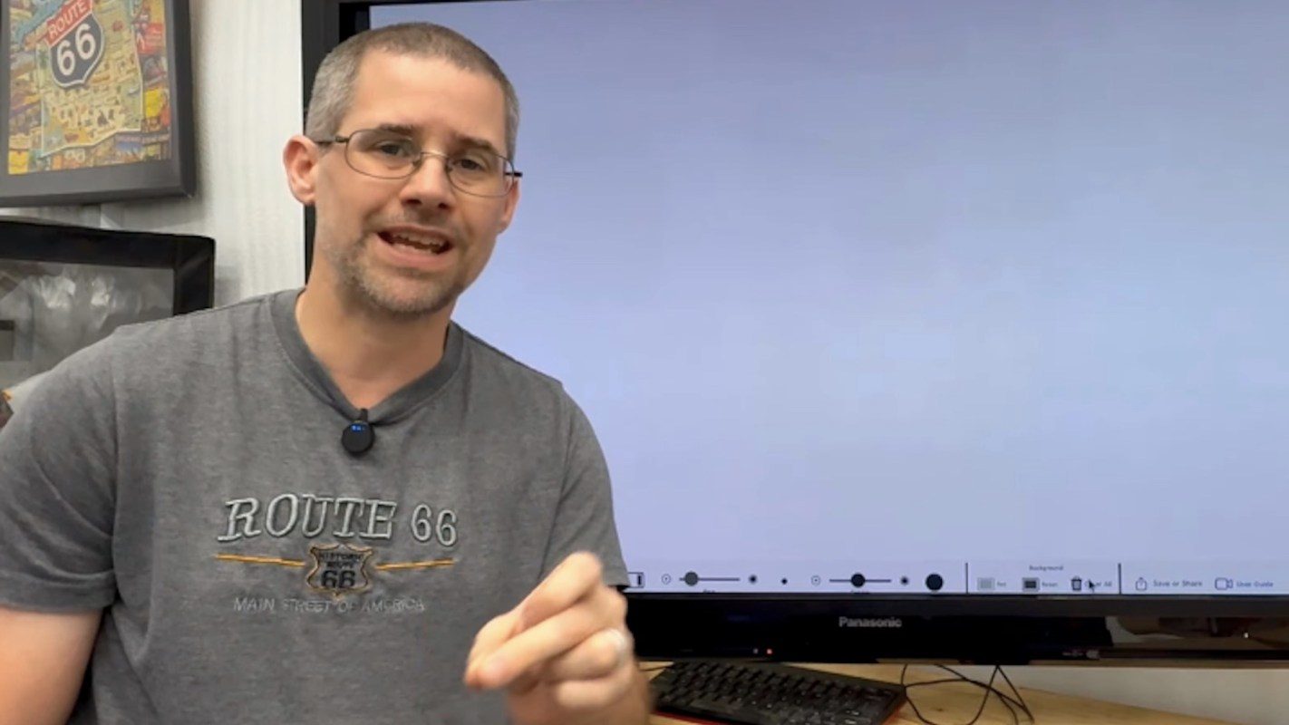 A man speaking in front of a blank screen and a computer workstation.