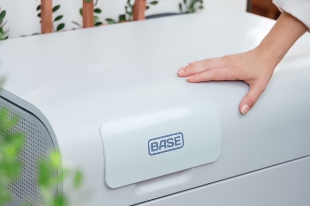 A hand rests on a gray device labeled "BASE" in a bright, plant-filled environment.