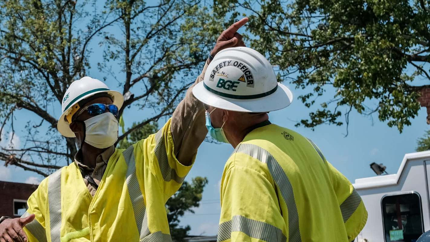 Two workers in safety gear discuss a project outdoors under a tree.
