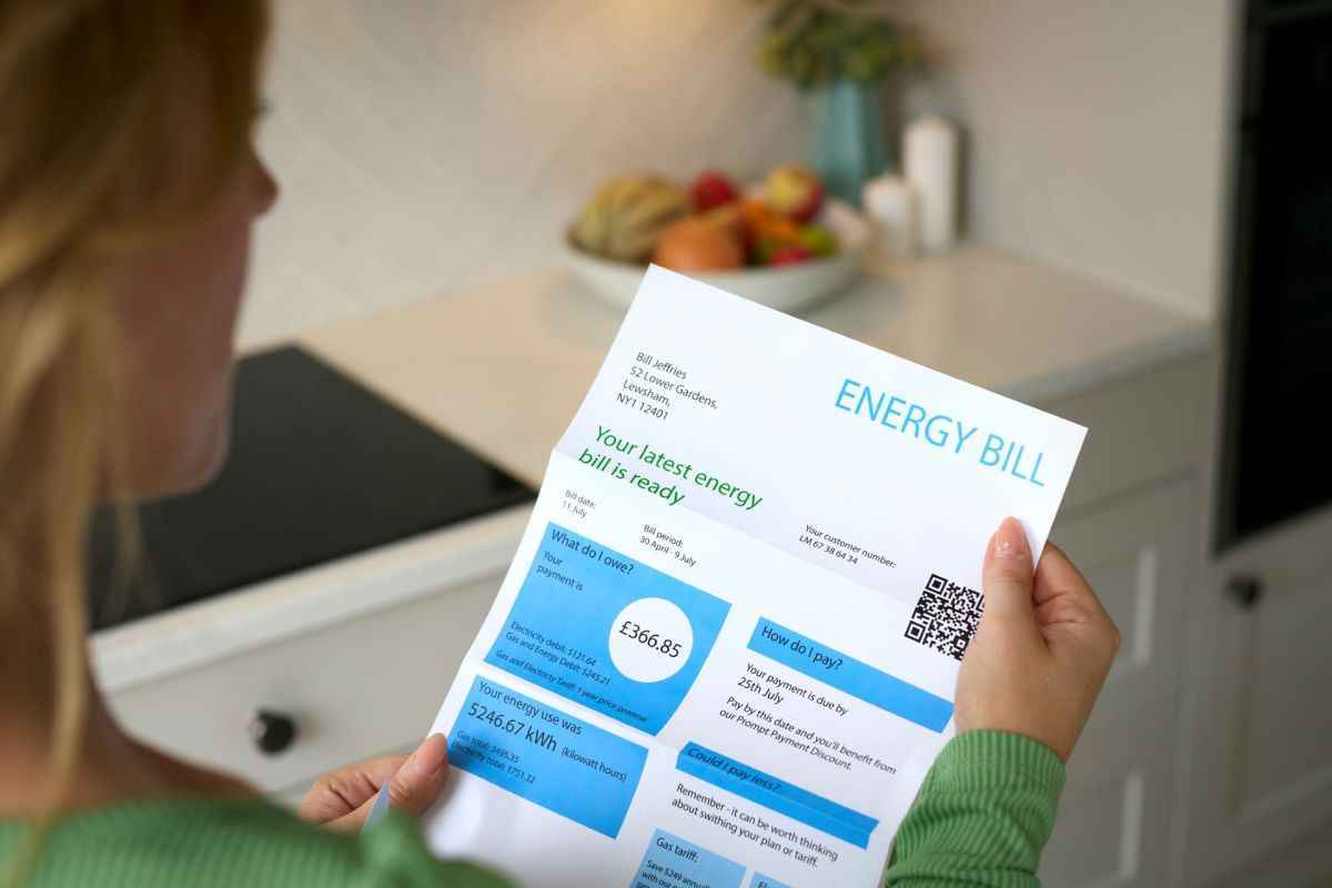 A person holds an energy bill in a kitchen, with fruits in a bowl in the background.