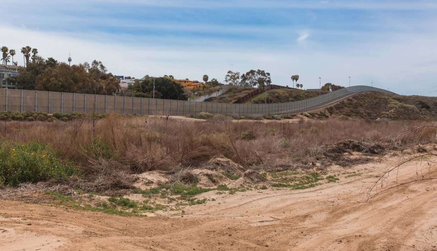 A border wall stretches across a hilly landscape with sparse vegetation under a cloudy sky.
