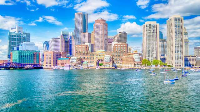 A city skyline with modern buildings along a waterfront, featuring boats in the water and blue skies.