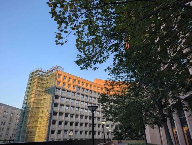 A building under construction with scaffolding, surrounded by trees and a clear sky.