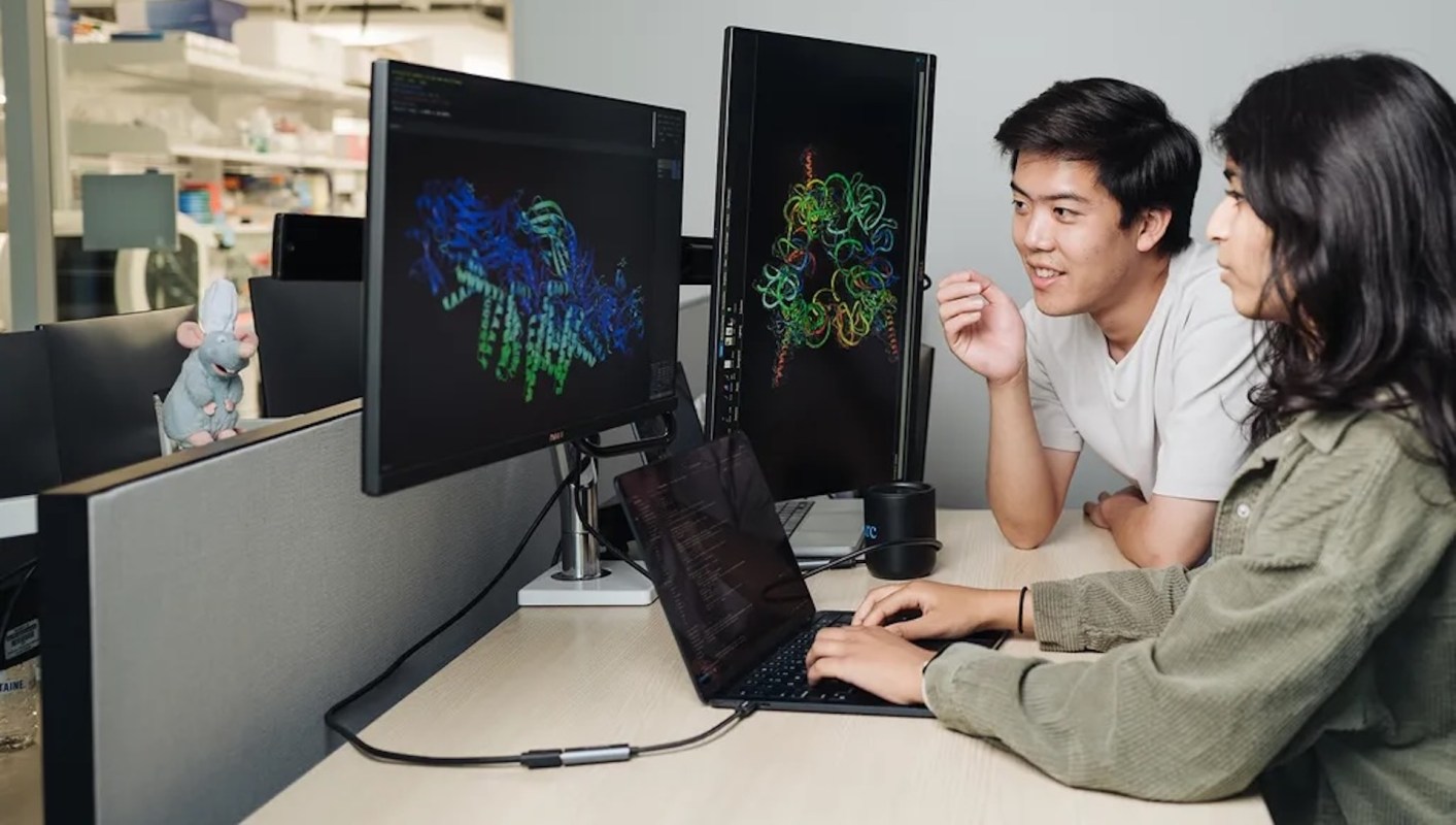 Two people work together at a desk with laptops and monitors displaying colorful data visualizations, with a toy mouse nearby.