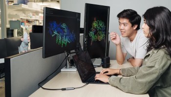 Two people work together at a desk with laptops and monitors displaying colorful data visualizations, with a toy mouse nearby.