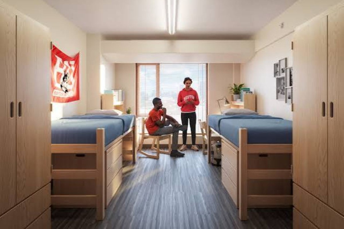 Two students in a dorm room with two beds and a window, engaged in conversation.