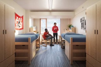 Two students in a dorm room with two beds and a window, engaged in conversation.