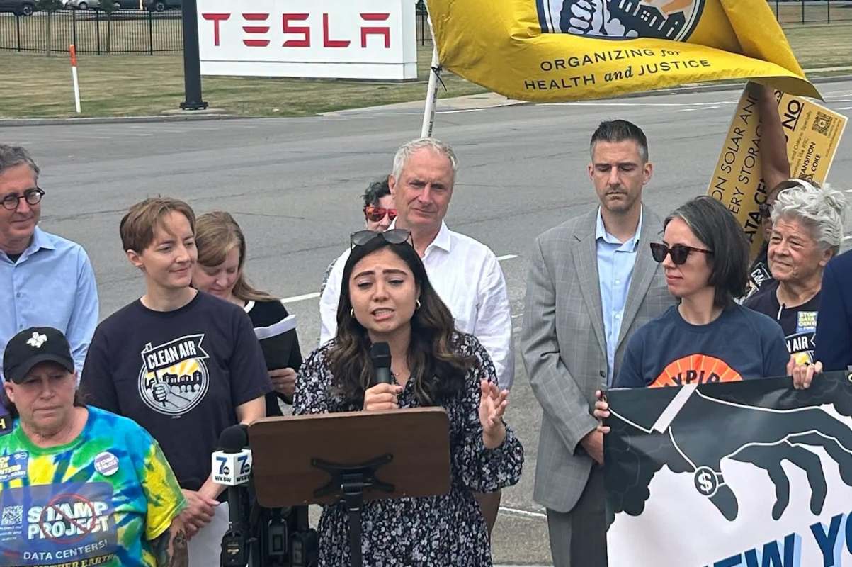 A group of people at a rally in front of a Tesla sign, with one person speaking into a microphone and holding a sign.
