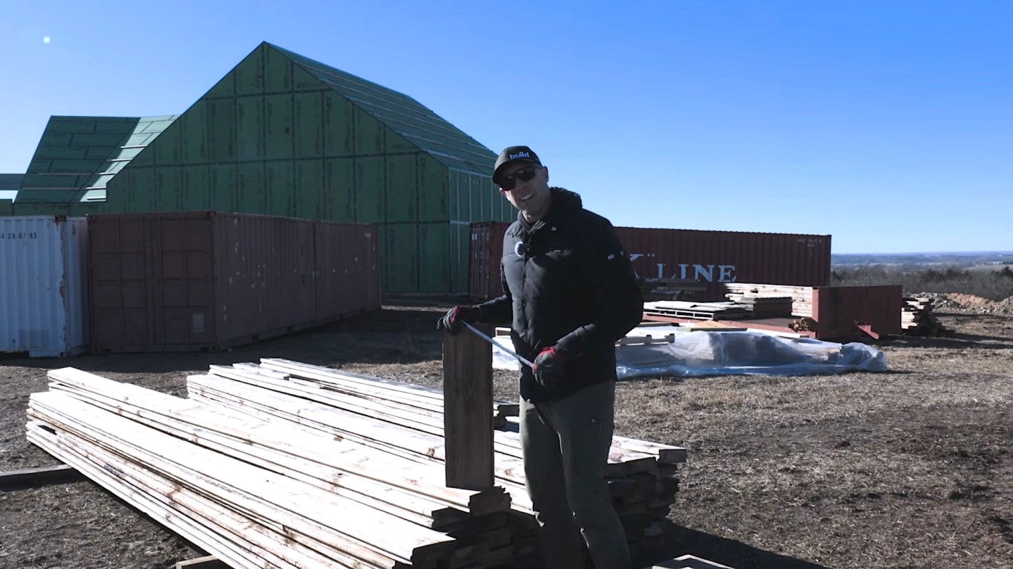 A person stands near stacks of wooden planks and shipping containers on a construction site.