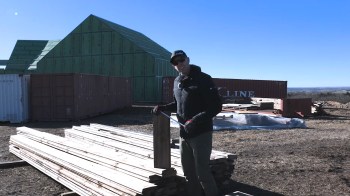 A person stands near stacks of wooden planks and shipping containers on a construction site.