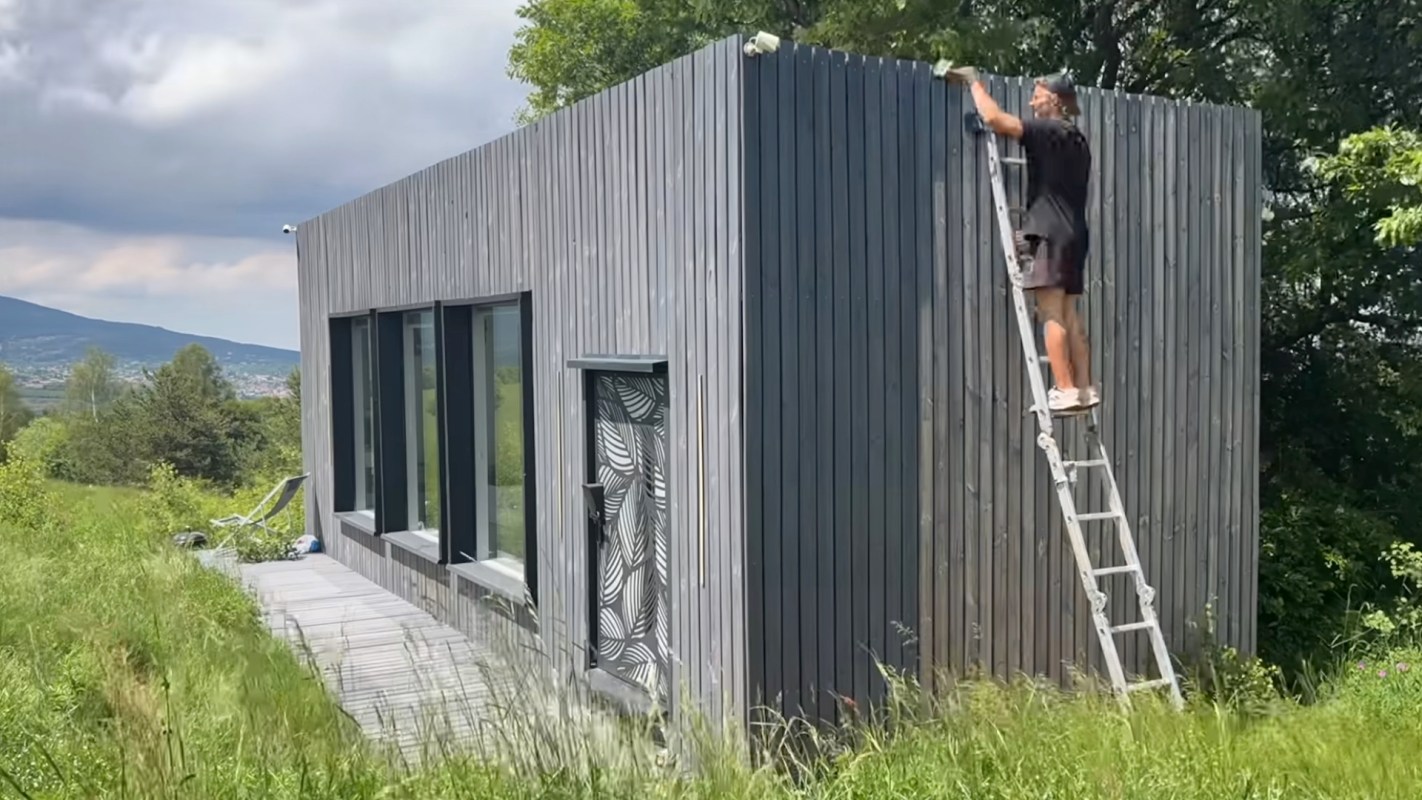 A person on a ladder works on the exterior of a modern wooden house surrounded by grass and trees.