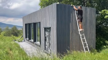 A person on a ladder works on the exterior of a modern wooden house surrounded by grass and trees.