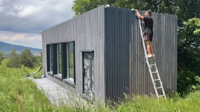 A person on a ladder works on the exterior of a modern wooden house surrounded by grass and trees.