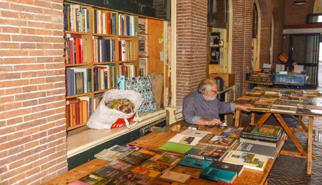 An elderly man examines books on a table in a room with a brick wall and shelves lined with books.