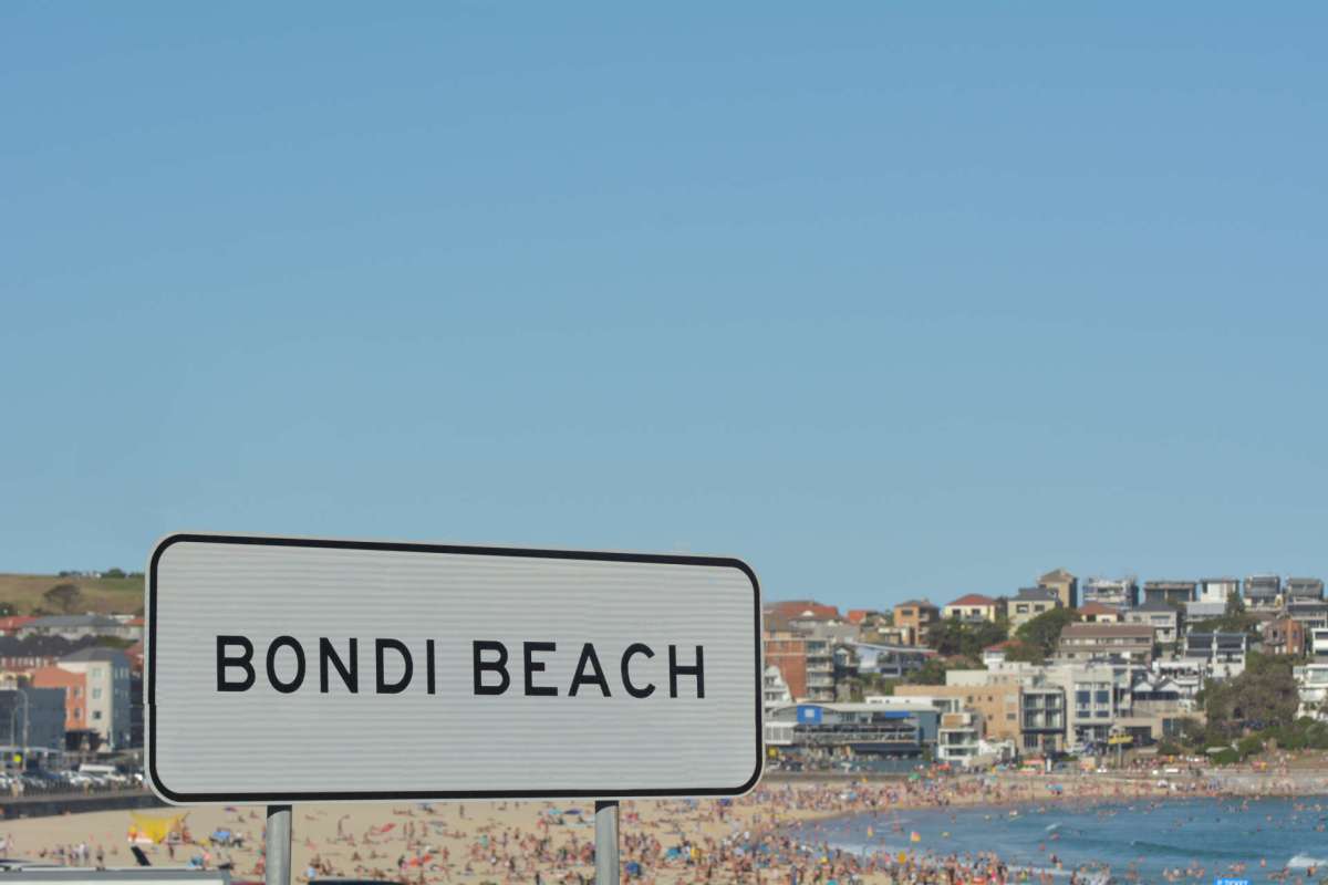 A sign for Bondi Beach with a beach and crowd in the background.