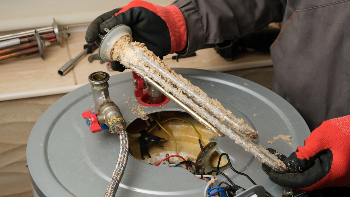 A person holds a corroded heating element above an open water heater tank.