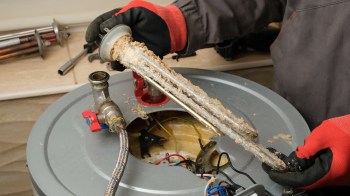 A person holds a corroded heating element above an open water heater tank.