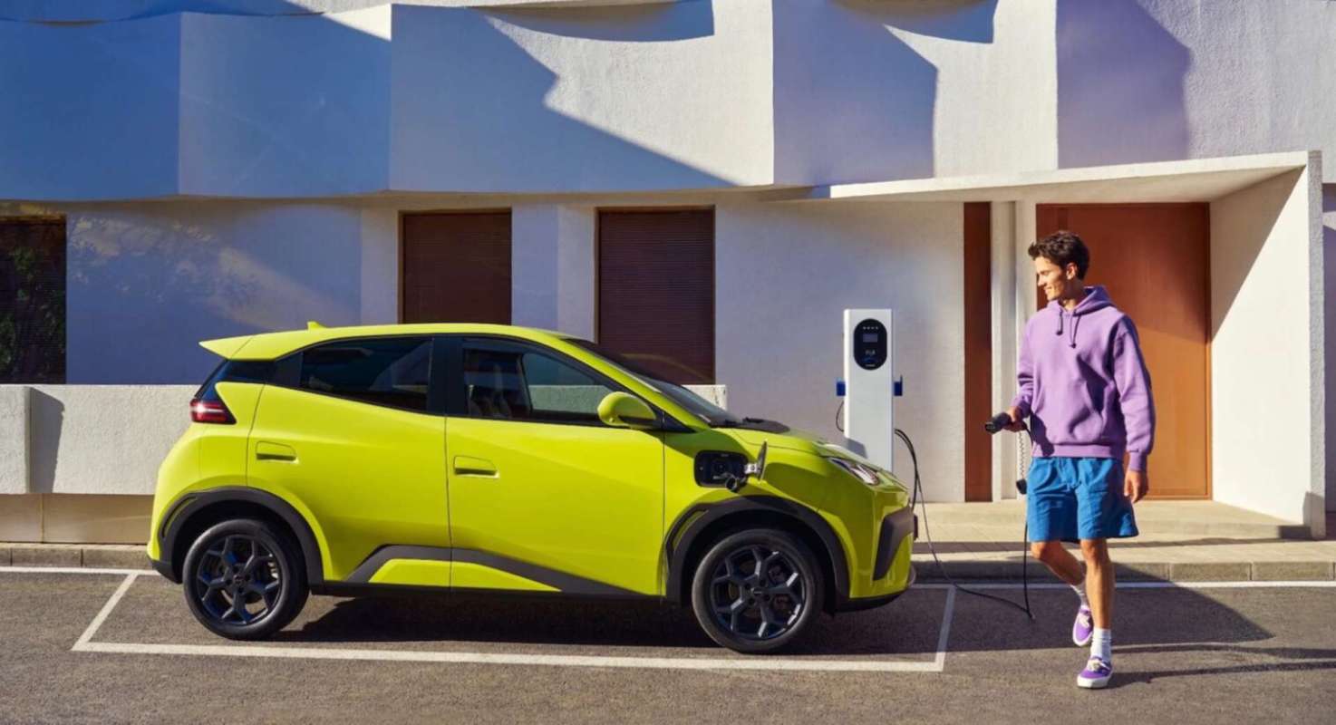 A man walks near an electric car connected to a charging station outside a modern building.