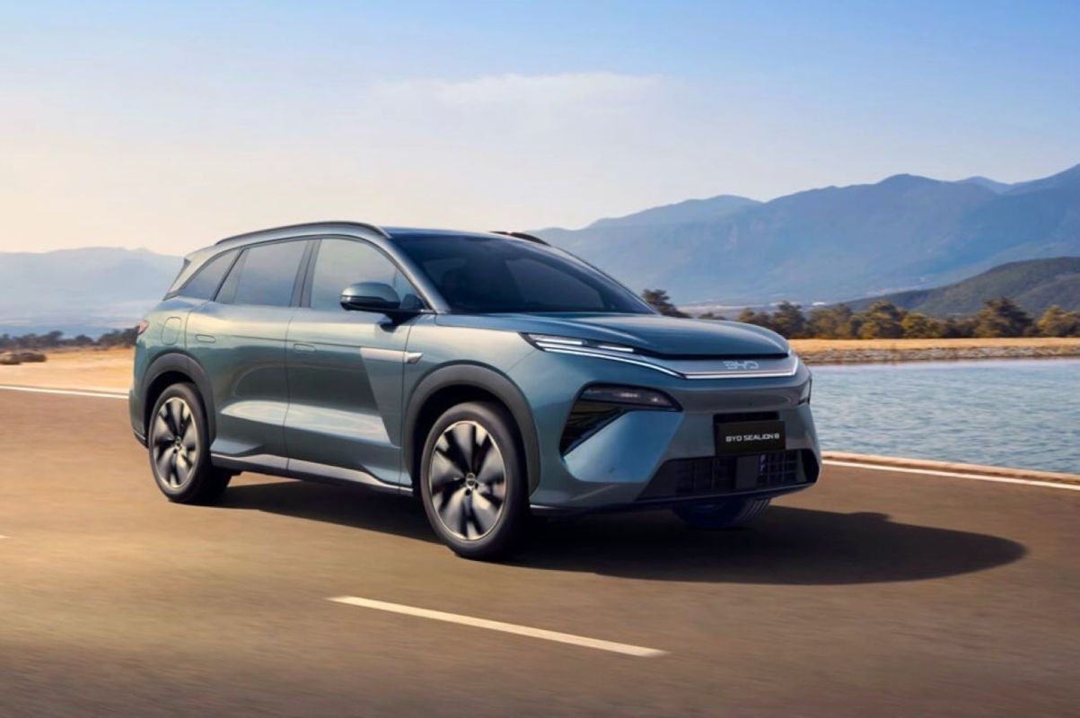 An electric SUV driving on a road beside a body of water with mountains in the background.
