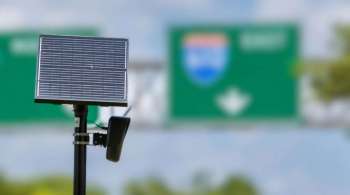 A solar panel mounted on a pole with highway signs visible in the background.