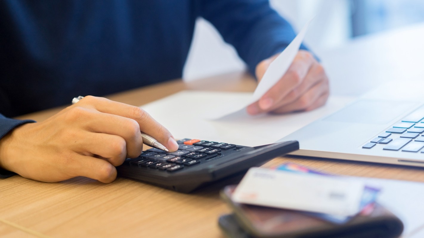 A person uses a calculator and holds a receipt while sitting at a desk with a laptop and cards.