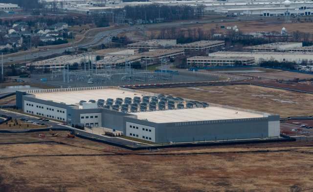 A large industrial building with rooftop equipment, surrounded by open land and distant structures.