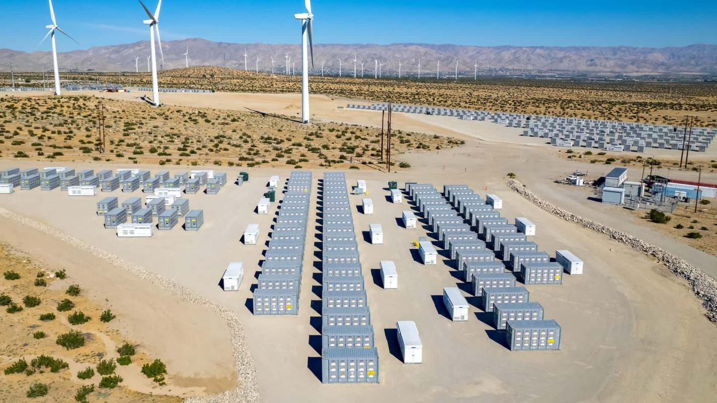 A grid of energy storage containers in a desert landscape with wind turbines in the background.