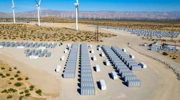 A grid of energy storage containers in a desert landscape with wind turbines in the background.