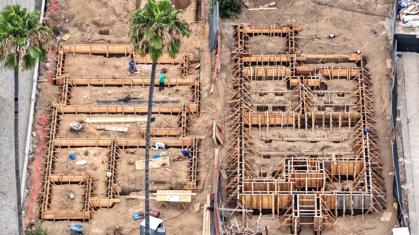 Construction site with wooden frames and workers preparing a foundation, surrounded by palm trees and construction materials.