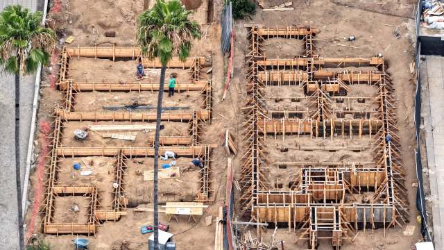 Construction site with wooden frames and workers preparing a foundation, surrounded by palm trees and construction materials.