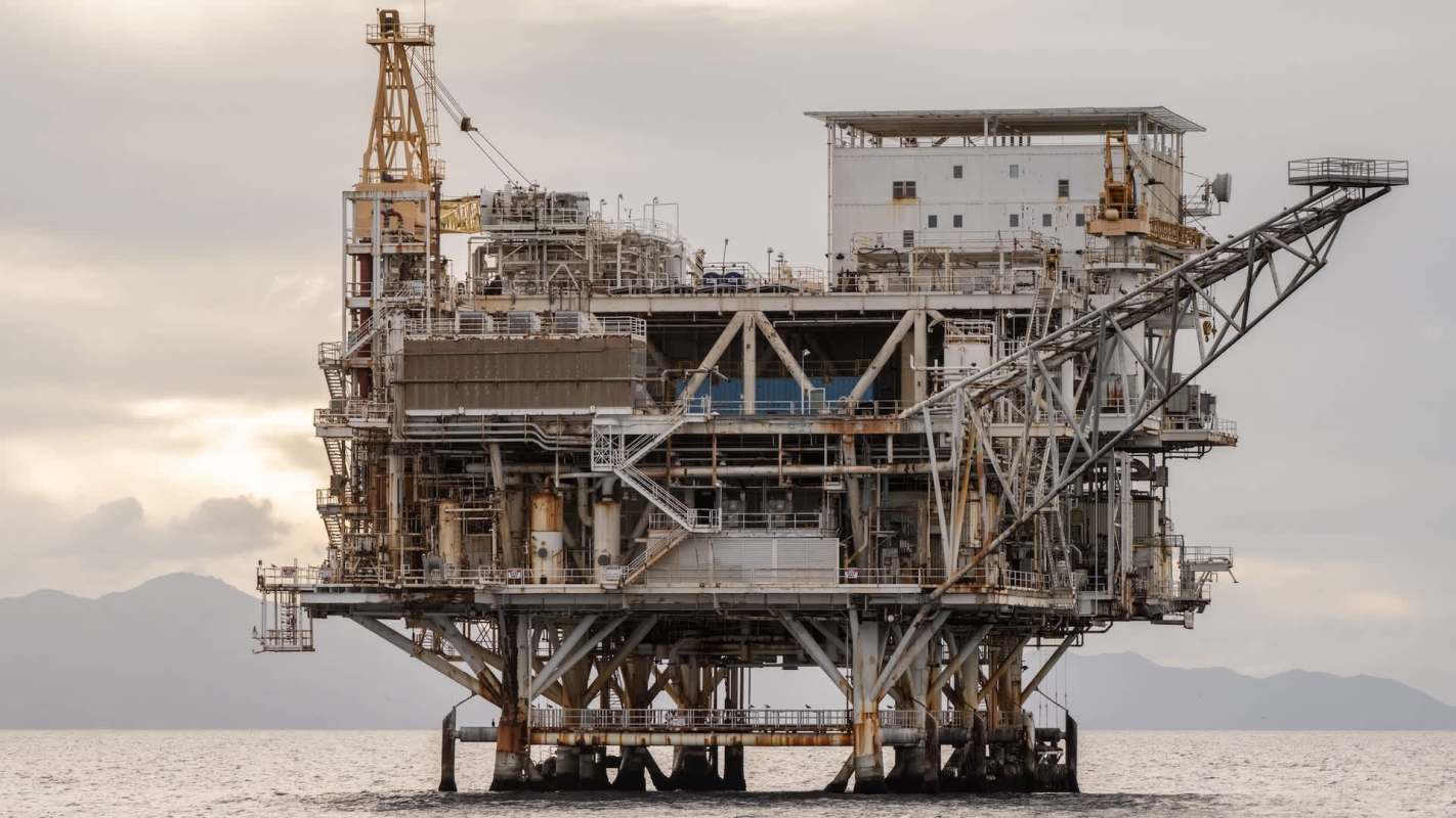 An oil drilling platform stands in the ocean with hills visible in the background.