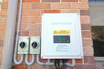 A solar inverter mounted on a brick wall with two adjacent DC isolators and warning labels.