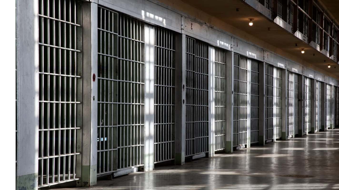 A row of prison cell doors with bars in a dimly lit corridor.
