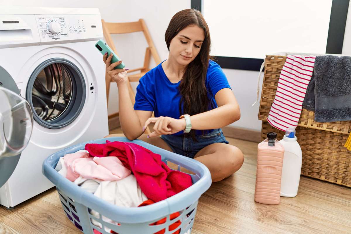 A woman checks her smartwatch while sitting next to a laundry basket and a washing machine.