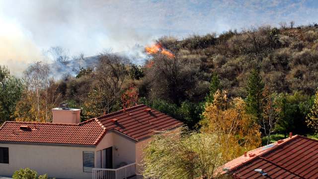 Flames and smoke rise from a hillside near residential houses surrounded by trees and shrubs.