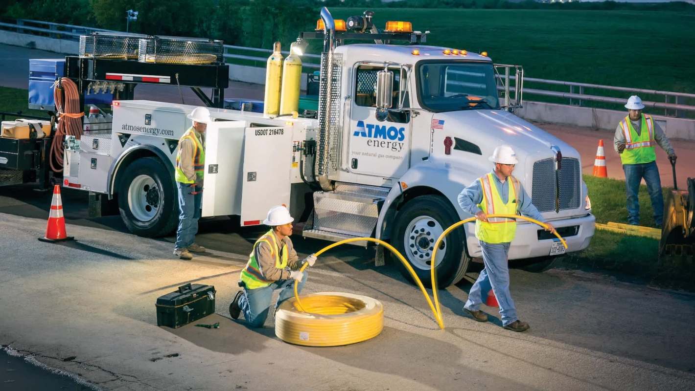 Four workers in safety gear handle equipment near an Atmos Energy service truck on a roadway.