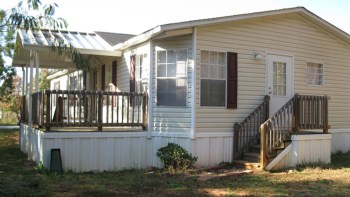 A manufactured mobile home with a porch and steps, surrounded by grassy areas and trees.
