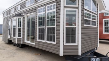 A modern tiny home with multiple windows and a light gray exterior, positioned on a trailer.