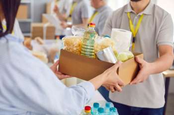 A person hands a box filled with food items to another person in a communal setting.