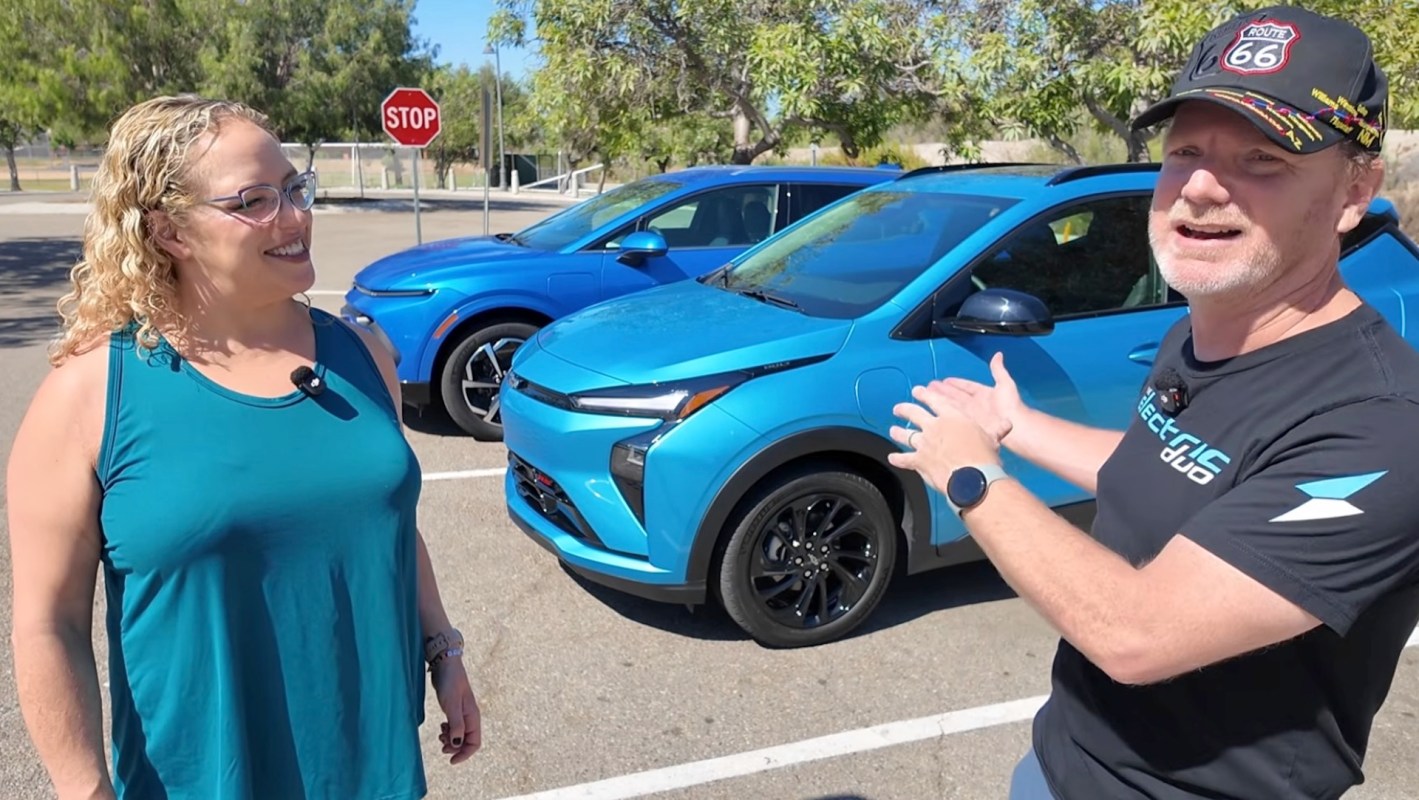 A man and a woman stand next to two blue electric cars in a parking lot.